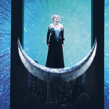 Scene from The Magic Flute by Wolfgang Amadeus Mozart: The Queen of the Night stands on a silver crescent moon in a sparkling dark gown, set against a radiant, star-like backdrop; her presence appears majestic and powerful.