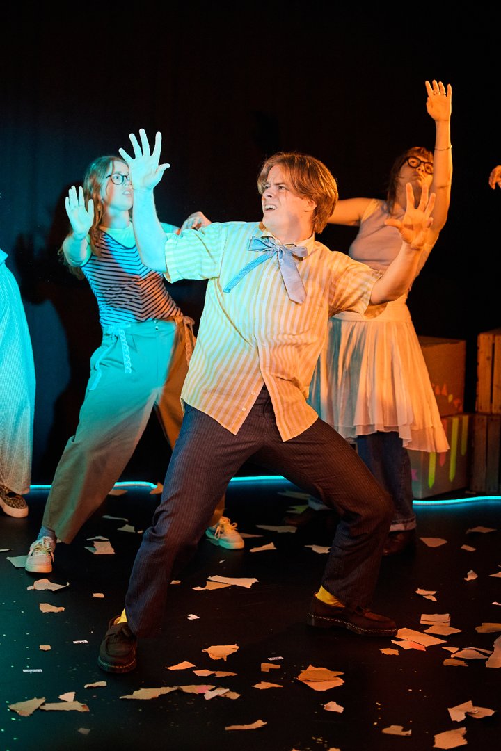 Szene aus When I find myself in Times of Trouble des Spielclubs Opera, Song and Scene. Mehrere Mitwirkende befinden sich in einer dynamischen Choreografie auf einer dunkel gehaltenen Bühne. Im Mittelpunkt steht eine Person in einem weiß und gelb gestreiften Hemd mit großer Schleife am Hals. Mit weit gespreizten Beinen und erhobenen Händen nimmt sie eine ausdrucksstarke Haltung ein, als würde sie sich gegen eine unsichtbare Kraft stemmen oder auf etwas Unerwartetes reagieren. Der Gesichtsausdruck wirkt angespannt und entschlossen zugleich. Hinter und neben ihr spiegeln weitere Personen ähnliche Bewegungen: Arme sind ausgestreckt, Hände geöffnet, die Körper in unterschiedliche Richtungen geneigt. Die Bühne ist in blaues und warmes orangefarbenes Licht getaucht, wodurch starke Kontraste entstehen. Auf dem Boden liegen zahlreiche verstreute Papierfetzen, die den Eindruck von Unordnung oder einem bereits stattgefundenen Ereignis vermitteln.