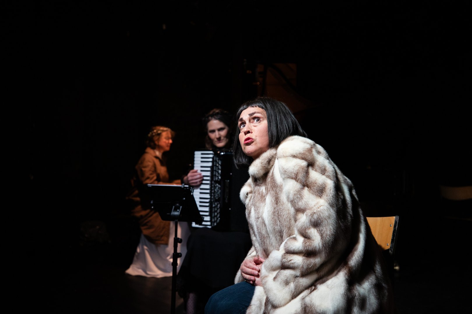 KS Dalia Schaechter in einem auffälligen Pelzmantel sitzt seitlich auf einem Stuhl im Vordergrund und blickt mit ausdrucksstarker Mimik nach oben. Im Hintergrund begleiten die Akkordeonspielerin Dorrit Bauerecker und die Pianistin Theresia Renelt die Szene. Die Bühne ist dunkel, mit gezieltem Licht auf die Darstellenden, was eine intime, dramatische Atmosphäre erzeugt.