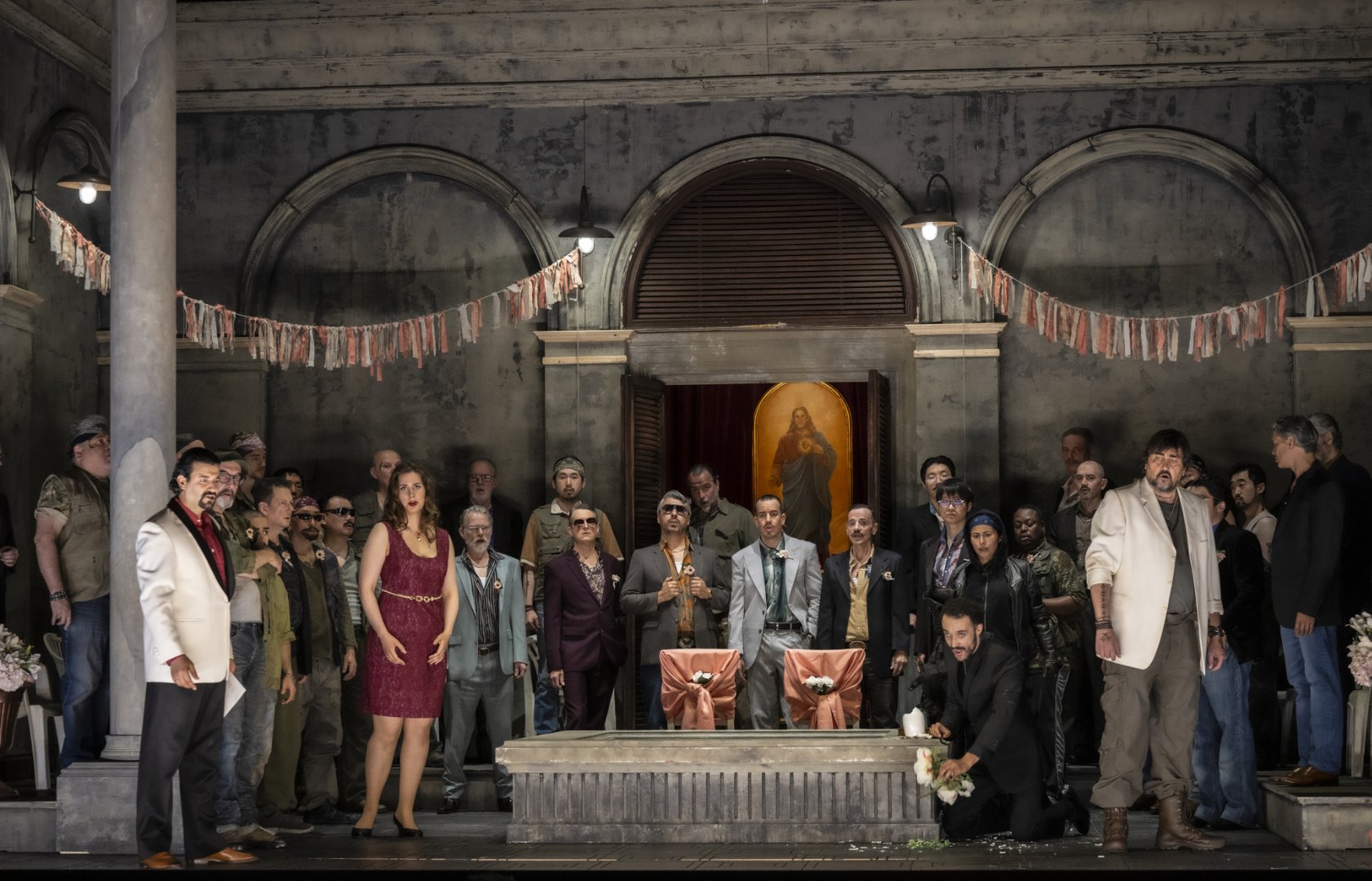 Auf der Bühne versammeln sich Argirio, Isaura, Roggiero, Tancredi, Orbazzano und der Chor der Oper Köln zu einer großen Ensembleszene aus Rossinis Tancredi. Die Figuren stehen vor der Fassade einer Villa mit Rundbögen und religiösem Bildnis im Hintergrund. In der Mitte befinden sich festlich geschmückte Stühle und Blumenschmuck, die auf eine Zeremonie oder Feier hinweisen. Während einige Figuren angespannt nach vorne blicken, kniet eine Person mit Blumen am Bühnenrand. Tancredi steht in dunkler Kleidung etwas abseits der Gruppe.