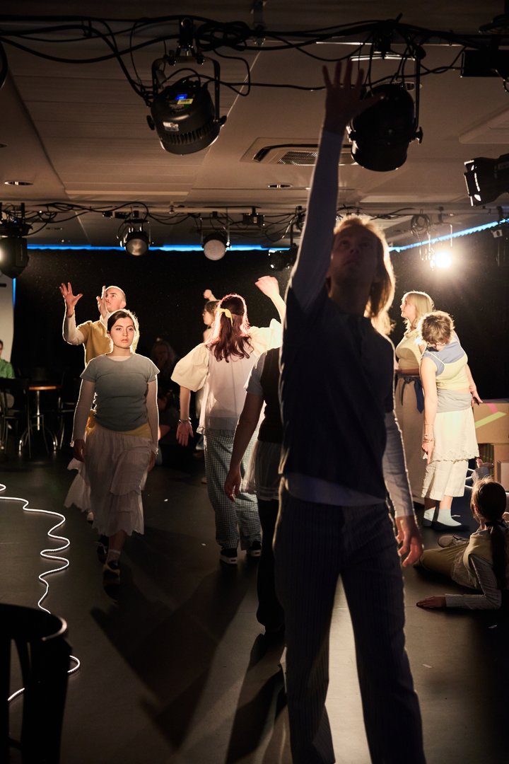 Szene aus When I find myself in Times of Trouble des Spielclubs Opera, Song and Scene. In einem offenen Bühnenraum bewegen sich zahlreiche Mitwirkende gleichzeitig durch den Raum. Im Vordergrund steht eine Person mit ausgestrecktem Arm, die ihre Hand weit nach oben richtet, als wolle sie nach etwas greifen oder einem Ziel entgegenstreben. Im Hintergrund sind weitere Personen in unterschiedlichen Bewegungen und Haltungen zu sehen: Einige gehen entschlossen voran, andere wenden sich voneinander ab, heben die Arme, verharren in Gedanken oder sitzen beziehungsweise liegen auf dem Boden. Die Bühne wird von starken Lichtquellen erhellt, die einzelne Figuren hervorheben und lange Schatten über den Boden werfen. Sichtbar sind auch die Scheinwerfer und technischen Elemente des Aufführungsraums, wodurch der Eindruck einer unmittelbaren, gemeinschaftlichen Performance entsteht.
Die Szene wirkt wie ein vielstimmiges Geflecht aus individuellen Wegen und Erfahrungen. Jede Person scheint einer eigenen Bewegung oder einem eigenen Impuls zu folgen, während sich die unterschiedlichen Handlungen zu einem gemeinsamen Bühnenbild verbinden. Der Raum ist von Dynamik geprägt: Menschen kommen zusammen, entfernen sich voneinander, suchen Orientierung oder richten ihren Blick nach oben und in die Ferne.