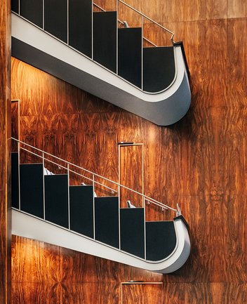 Zwei elegant geschwungene Ränge im Saal der Oper Köln am Offenbachplatz mit dunklen Brüstungen und hellen Handläufen. Die Wände sind vollständig mit warm schimmernder Holzvertäfelung verkleidet, die durch indirektes Licht besonders zur Geltung kommt und dem Raum eine ruhige, hochwertige Atmosphäre verleiht.