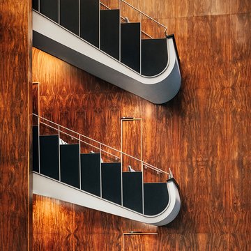 Zwei elegant geschwungene Ränge im Saal der Oper Köln am Offenbachplatz mit dunklen Brüstungen und hellen Handläufen. Die Wände sind vollständig mit warm schimmernder Holzvertäfelung verkleidet, die durch indirektes Licht besonders zur Geltung kommt und dem Raum eine ruhige, hochwertige Atmosphäre verleiht.