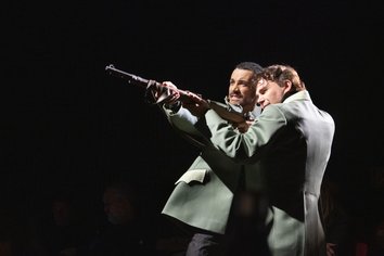 Zwei Darsteller verkörpern gemeinsam Max aus der Oper „Freikugeln“ und stehen eng nebeneinander auf einer dunklen Bühne. Beide tragen grüne Jacken. Sie halten zusammen ein Gewehr, das nach links oben gerichtet ist, wobei der vordere Darsteller zielt und angespannt blickt, während der hintere ihn von der Seite unterstützt und sich dicht an ihn lehnt. Ihre Körper sind leicht nach vorne geneigt, die Haltung wirkt konzentriert und spannungsvoll. Das Licht hebt die beiden Figuren stark aus dem dunklen Hintergrund hervor und lenkt den Fokus auf die gemeinsame Handlung.