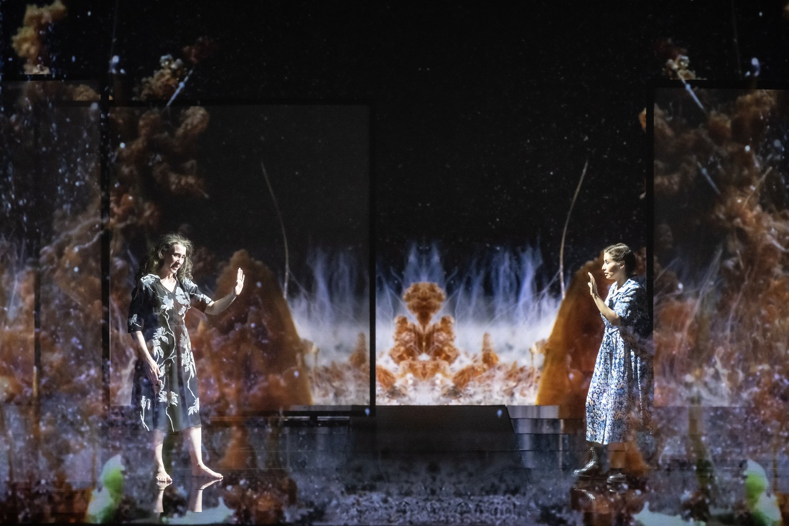 Zabelle und die Woman stehen sich in einer weitläufigen Bühnenlandschaft aus der Oper Picture a day like this gegenüber. Beide Figuren sind klein vor einer monumentalen Projektion positioniert, die abstrakte, wolkenartige Formen in Blau-, Weiß- und Rosttönen zeigt und an kosmische oder unterwasserartige Bilder erinnert. Die dunkle Bühne spiegelt Licht und Farben der Projektion wider und verstärkt die traumartige Wirkung der Szene. Die beiden Frauen stehen ruhig und einander zugewandt im Zentrum der eindrucksvollen visuellen Komposition.