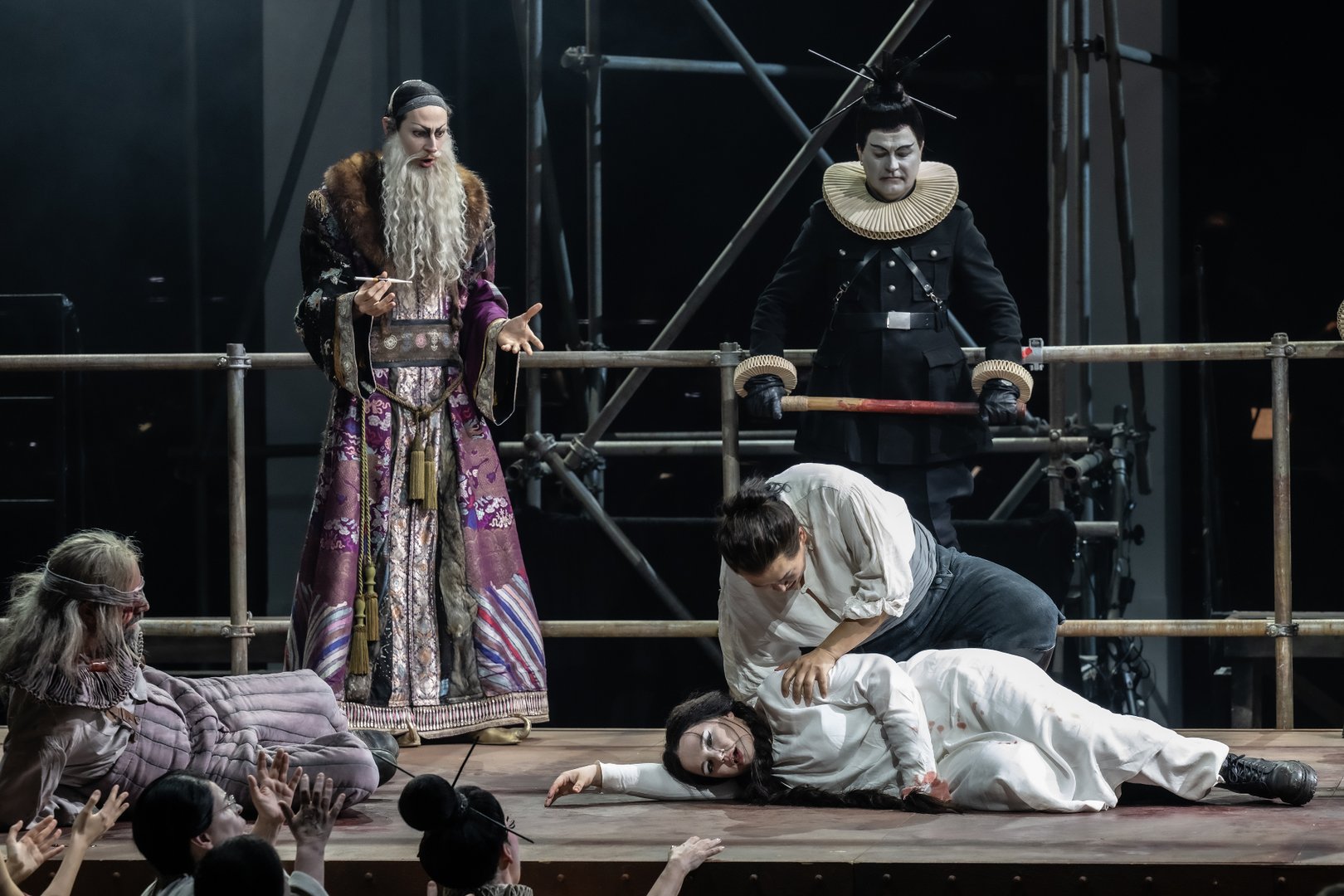 Zu sehen sind Timur, ein Mandarin, ein Chormitglied der Oper Köln, Calaf und Liù in einer dramatischen Szene aus „Turandot“. Im Vordergrund liegt Liù in weißer Kleidung reglos am Boden, während Calaf sich verzweifelt über sie beugt. Links sitzt der blinde Timur erschüttert neben der Szene. Hinter den Figuren stehen ein Mandarin in prunkvollem Gewand sowie ein Chormitglied in schwarzem Kostüm mit großer Halskrause. Das kühle Licht und die Gerüstkonstruktionen im Hintergrund verstärken die düstere und emotionale Atmosphäre der Inszenierung.