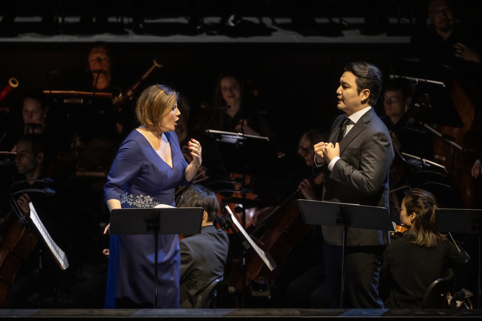 Auf der Bühne stehen Marta Torbidoni (Elvira) und Insik Choi (Don Carlos) einander gegenüber und singen mit ausdrucksstarker Gestik, jeweils an eigenen Notenpulten. Im Hintergrund ist das Gürzenich Orchester zu sehen, dessen Musiker*innen konzentriert ihre Instrumente spielen und die Szene musikalisch begleiten.