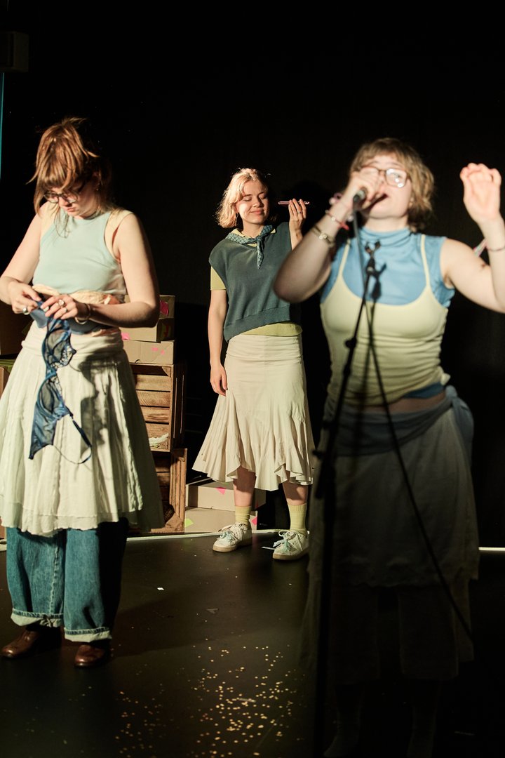Szene aus When I find myself in Times of Trouble des Spielclubs Opera, Song and Scene. Drei Mitwirkende befinden sich auf einer dunkel gehaltenen Bühne, die von warmem Licht punktuell erhellt wird. Im Vordergrund rechts singt eine Person in ein Mikrofon. Mit erhobener Hand und geöffnetem Mund wirkt sie ganz in den Moment vertieft. Links steht eine weitere Person, die konzentriert einen blauen BH in den Händen hält und ihren Blick darauf richtet. In der Bildmitte steht eine dritte Person mit geschlossenen Augen und einem leichten Lächeln. Sie hält ein Smartphone in der Hand an ihr Ohr und scheint aufmerksam zuzuhören oder in Gedanken versunken zu sein. Hinter ihr sind Holzkisten und Kartons als Teil des Bühnenbilds zu erkennen. Auf dem Boden liegen verstreute kleine helle Partikel, die im Licht glitzern.