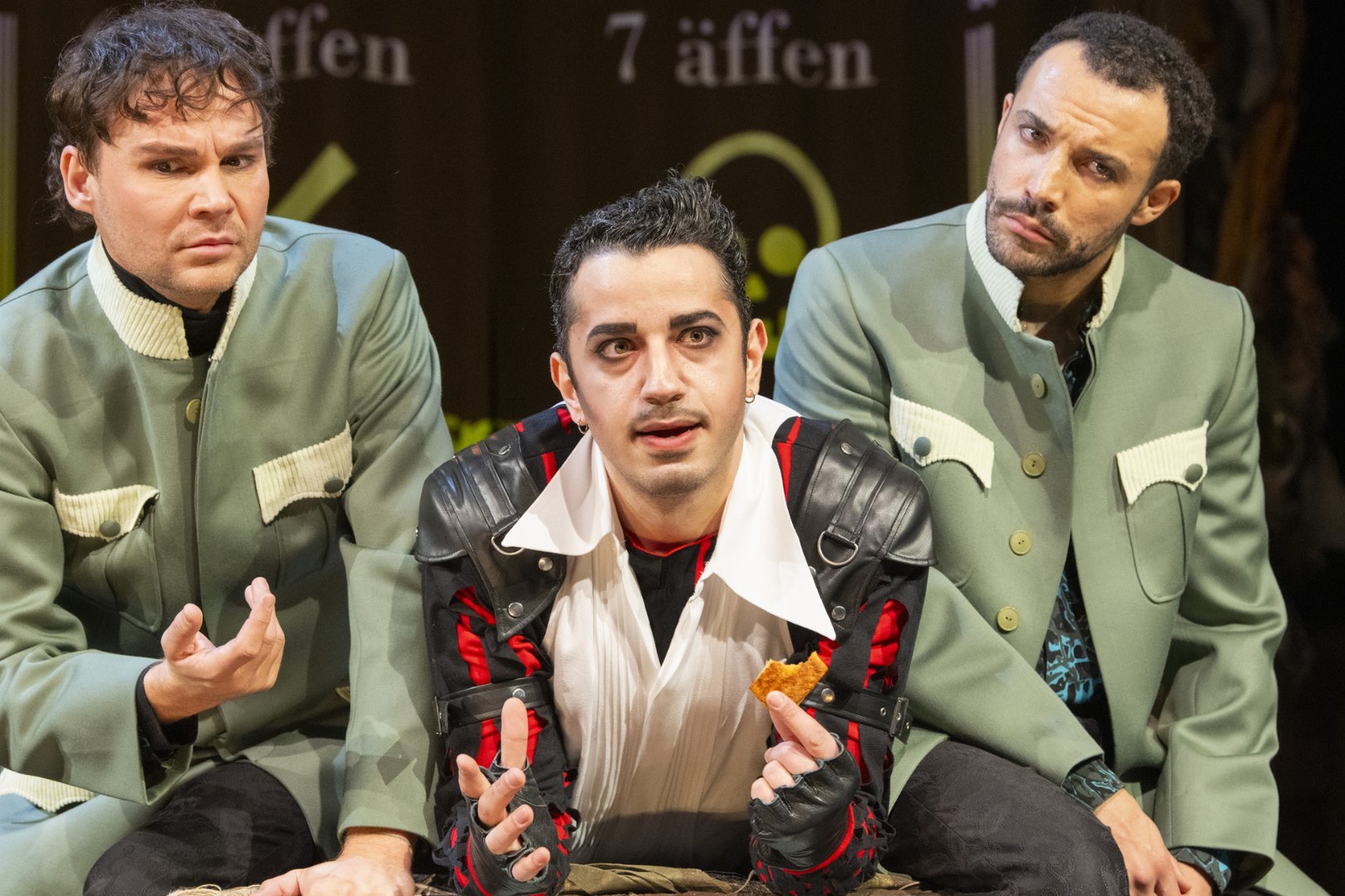 Drei Darsteller sitzen dicht nebeneinander auf einer Theaterbühne und blicken aufmerksam nach vorne. In der Mitte sitzt Kaspar, erkennbar an seinem auffälligen Kostüm mit schwarzer Lederjacke, roten Akzenten und einem weißen Hemd. Er gestikuliert intensiv mit beiden Händen und hält ein kleines Stück Brot in der rechten Hand. Sein Gesichtsausdruck wirkt angespannt und eindringlich.
Links und rechts neben ihm sitzen zwei Darsteller der Figur Max in nahezu identischen, grünlichen Kostümen mit hellen Einsätzen. Beide reagieren auf Kaspar und wirken konzentriert und leicht irritiert. Der Darsteller rechts nutzt Deutsche Gebärdensprache (DGS) und macht die Figur Max somit zweisprachig erfahrbar.
Im Hintergrund sind schemenhaft Schriftzüge zu erkennen, darunter „7 äffen“, sowie grafische Elemente, die an eine stilisierte Tafel erinnern. Die Szene stammt aus der Oper Freikugeln und zeigt eine intensive Interaktion zwischen den Figuren Max und Kaspar.