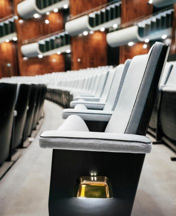 Seitliche Nahaufnahme einer Sitzreihe im Opernsaal der Oper Köln am Offenbachplatz mit gepolsterten Sitzen und integrierten Platznummern. Die Perspektive entlang der Reihe betont die Tiefe des Raums und die hochwertige Ausstattung.