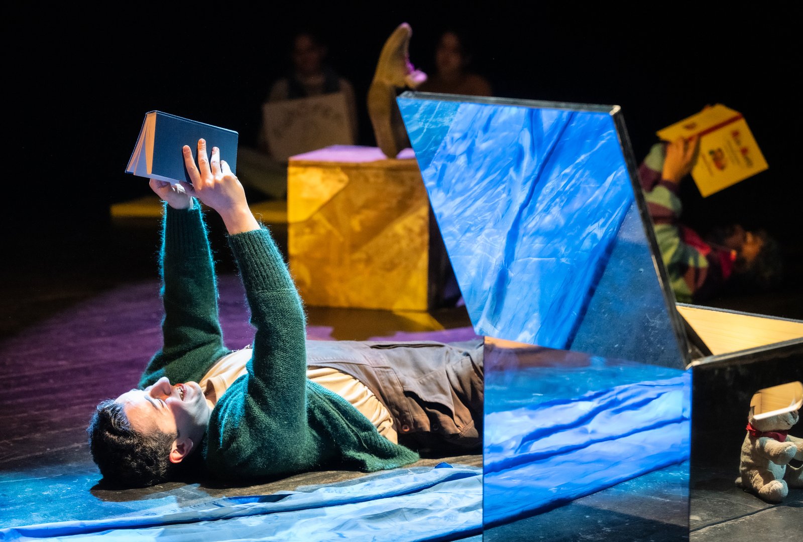 Szene aus der Kinderoper Max und Moritz: Auf einer dunklen Bühne liegt Henriette, das Huhn / Herr Guru / Dr. Gehirn / Belinda, die Tarantel. Der Darsteller trägt eine dunkle Latzhose über einem hellen Shirt; darüber trägt er eine gemütliche grüne Strickjacke. Im Vordergrund steht ein Spiegelwürfel, in dem sich ein Stoff-Teddy-Bär spiegelt. Im Hintergrund sind weitere Personen liegend oder sitzend angedeutet.