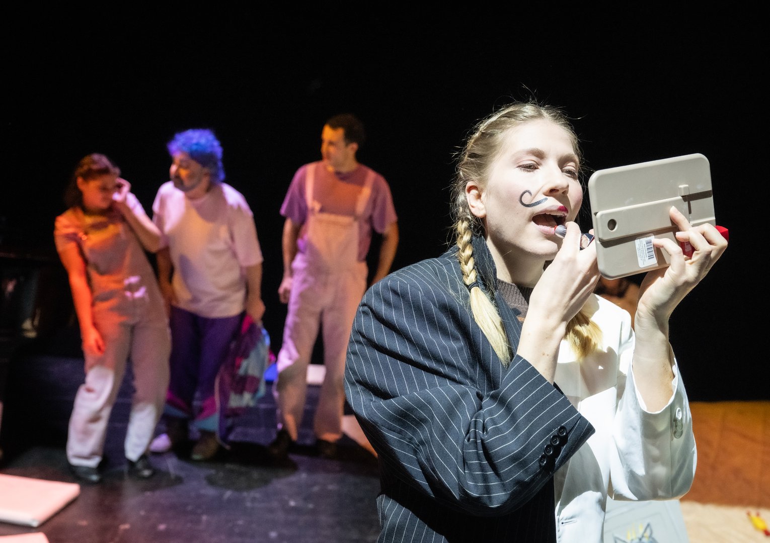 Szene aus der Kinderoper Max und Moritz: Im Vordergrund steht Henny, das Huhn / Die Helferin / Überglucke im Scheinwerferlicht und schminkt sich: Sie hält einen kleinen Spiegel in der Hand und trägt jeweils bis zur Hälfte der Lippen roten Lippenstift auf. Ihr Gesicht ist theatral geschminkt, inklusive eines aufgemalten Schnurrbarts. Sie trägt eine Jacke, die auf der einen Hälfte dunkel, gestreift und auf der anderen Seite weiß ist und an einen Anzug erinnert; ihre Haare sind zu zwei Zöpfen geflochten. Im Hintergrund sind Trouble mit blauer Lockenperücke und pinkem Oberteil und lilafarbenem Trainingsanzug sowie Max und Moritz in hellrosa Latzhose zu sehen, alle etwas unscharf vor dunklem Bühnenraum.