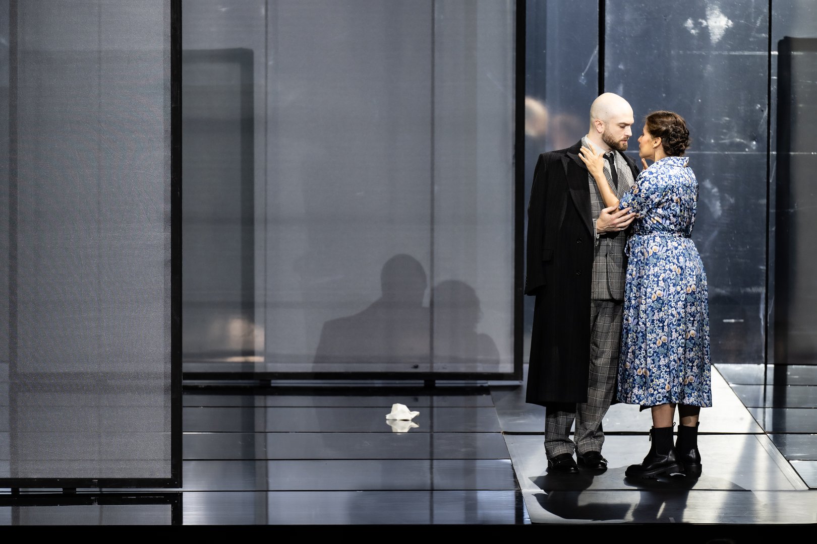 Zwei Figuren stehen sich auf einer modernen, dunkel gehaltenen Bühne nahe gegenüber: die Woman in einem blau geblümten Kleid und der Artisan / Collector im grauen Anzug mit langem schwarzem Mantel. Während sie sich intensiv ansehen und berühren, spiegeln sich ihre Silhouetten im glänzenden Bühnenboden und in den hohen, transparenten Wandelementen im Hintergrund. Die reduzierte Szenerie und das kalte Licht erzeugen eine intime, melancholische Stimmung.
