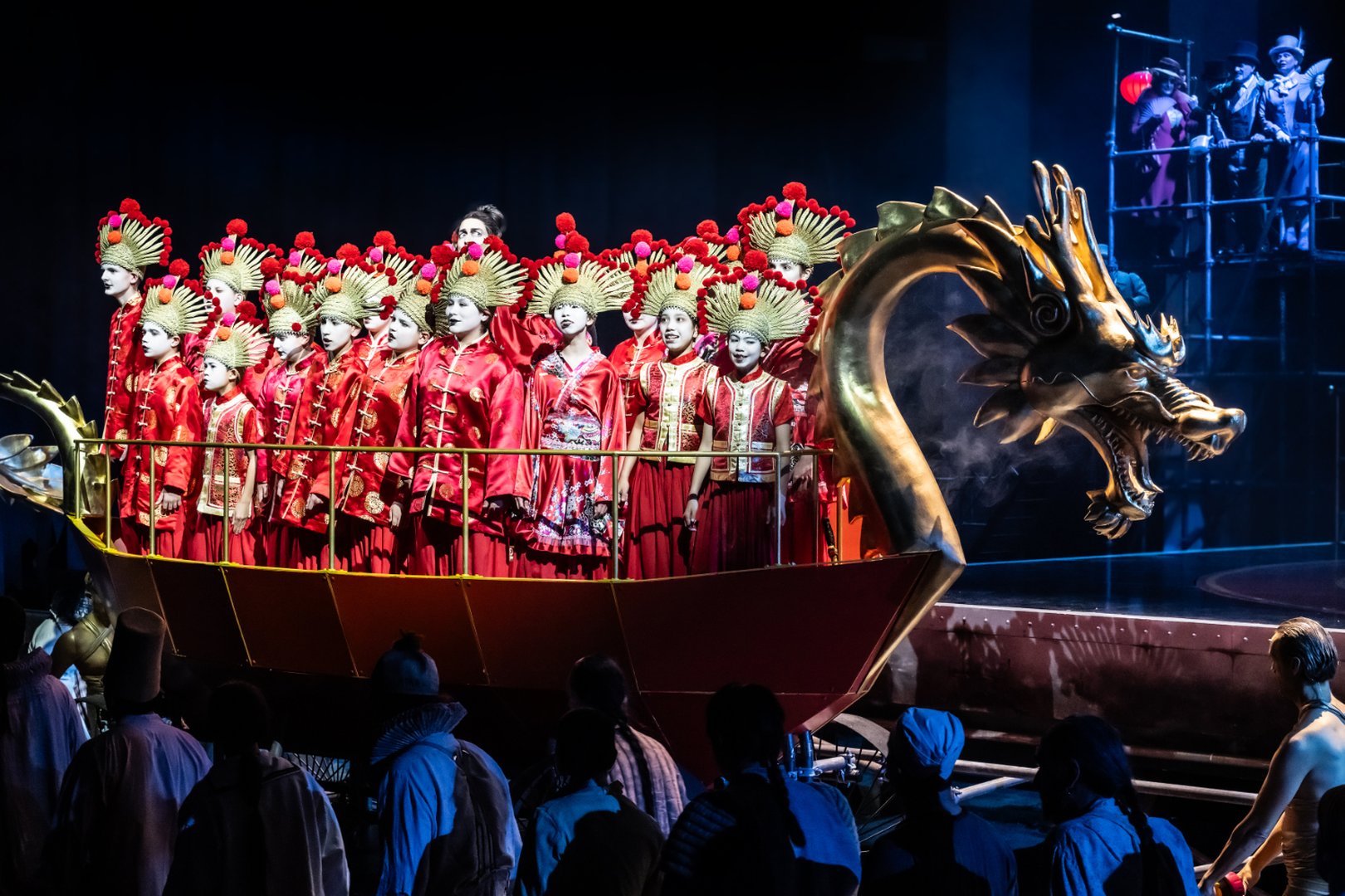 Zu sehen sind die Knaben des Kölner Domchors in einer Szene aus „Turandot“. Die Kinder tragen leuchtend rote Kostüme mit goldenen Kopfbedeckungen und stehen dicht gedrängt in einem stilisierten Drachenboot mit goldenem Drachenkopf. Die Bühne ist in dunkles Blau getaucht, wodurch die roten und goldenen Farben besonders hervorstechen. Im Hintergrund beobachten weitere Figuren das Geschehen von einer erhöhten Plattform.