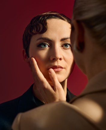 Nahaufnahme zweier Personen vor einem intensiven roten Hintergrund mit cineastischer Bildwirkung und warmer Lichtgebung: Im Zentrum steht eine androgyn wirkende Person mit kurzen, rot-braunen Haaren, die sorgfältig mit Wasserwelle gelegt sind. Ihre blauen Augen blicken ruhig und direkt nach vorn in Richtung des Betrachters. Ihr Gesicht ist weich ausgeleuchtet, die Haut wirkt glatt und ebenmäßig, der Ausdruck ist ernst, beinahe nachdenklich. Sie trägt ein schlichtes, dunkles Jackett, das ihre klare, reduzierte Erscheinung unterstreicht. Von rechts im Vordergrund ist eine zweite, sehr weiblich wirkende Person mit blonden, zum Zopf gebundenen Haaren nur teilweise sichtbar – unscharf, mit dem Rücken zur Kamera. Sie trägt einen hellen, beigefarbenen Blazer, der weich fällt und ihre Silhouette betont. Ihre Hand liegt sanft an der Wange der zentralen Figur, die Fingerspitzen berühren das Gesicht behutsam. Zwischen beiden entsteht eine intime, gespannte Nähe. Die Szene evoziert die Figuren aus der Oper Der Rosenkavalier von Richard Strauss, darunter die Feldmarschallin und entweder Octavian und Sophie, wobei die genaue Zuordnung bewusst offen bleibt.