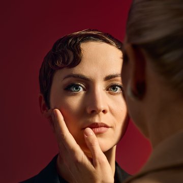 Close-up of two people against an intense red background with a cinematic look and warm lighting: At the center stands an androgynous-looking person with short, reddish-brown hair, carefully styled in finger waves. The persons blue eyes gaze calmly and directly forward toward the viewer. The face is softly lit, the skin appears smooth and even, and their expression is serious, almost contemplative. The person is wearing a simple, dark jacket that emphasizes the clean, understated appearance.
On the right in the foreground, a second, very feminine-presenting person with blonde hair tied back in a braid is only partially visible – out of focus, with the back to the camera. She is wearing a light beige blazer that falls softly and accentuates her silhouette. Her hand rests gently on the central figure’s cheek, with her fingertips touching the face with care. An intimate, charged closeness emerges between them. The scene evokes characters from the opera The Knight of the Rose by Richard Strauss, including the Marschallin and either Octavian and Sophie, with the exact interpretation intentionally left open.