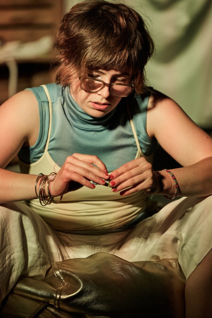 Szene aus When I find myself in Times of Trouble des Spielclubs Opera, Song and Scene. Eine Person sitzt allein auf dem Boden und ist ganz in eine kleine Tätigkeit vertieft. Mit gesenktem Blick hält sie einen Geldschein zwischen den Fingern und betrachtet ihn aufmerksam. Sie trägt eine Brille, mehrere Armbänder und helle Kleidung. Das warme Bühnenlicht fällt auf ihr Gesicht und ihre Hände, während der Hintergrund unscharf bleibt. Vor ihr liegt eine große dunkle Tasche oder ein Beutel, der einen Teil des Bildvordergrunds einnimmt. Die enge Bildkomposition schafft einen intimen Eindruck und lenkt die Aufmerksamkeit auf die konzentrierte Haltung der Person.