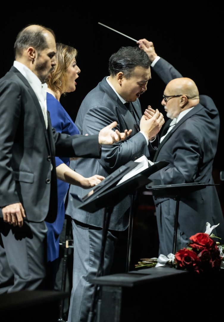 Auf der Bühne singen Adrian Sâmpetrean (Don Ruy Gómez de Silva), Marta Torbidoni (Elvira) und Young Woo Kim (Ernani) ausdrucksstark, versammelt um Notenpulte. Neben ihnen dirigiert Giuliano Carella (musikalische Leitung) mit erhobenem Taktstock und führt das Ensemble mit konzentrierter Intensität.