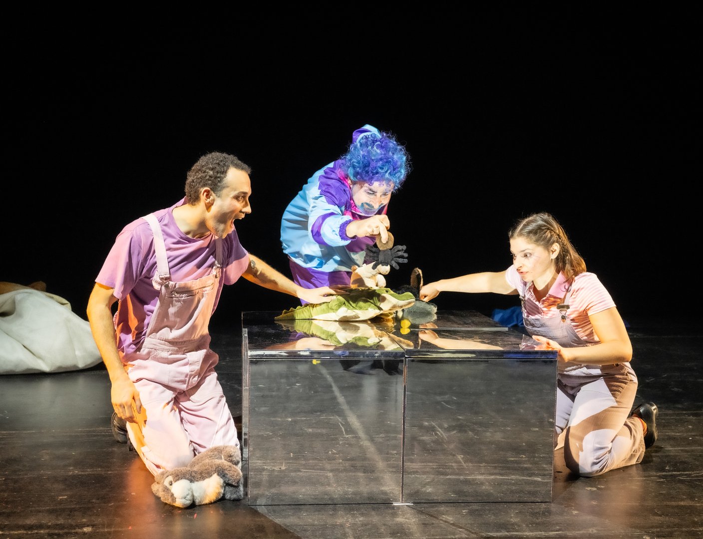 Szene aus der Kinderoper Max und Moritz: Vier Darsteller*innen in den Rollen von Henny, das Huhn / Die Helferin / Überglucke, Frau Gagackel, Henriette, das Huhn / Herr Guru / Dr. Gehirn / Belinda, die Tarantel und Trouble stehen in einer lebhaften Szene auf einer hellrosa beleuchteten Matte vor schwarzem Hintergrund. Drei Personen tragen dunkle Latzhosen über hellen Oberteilen; die vorderste Person trägt zusätzlich eine weiße Strickjacke und leuchtend rote Socken. Sie haben weiße Handschuhe mit orangenen Spitzen an den Händen, die wie Enten aussehen, und nehmen dynamische, fast tänzerische Posen ein. Rechts im Bild steht eine Person in einem hellblauen Retro-Trainingsanzug mit bunten Streifen und einer auffälligen, lockigen blauen Perücke. Alle Darsteller haben einen freudigen oder überraschten Gesichtsausdruck.