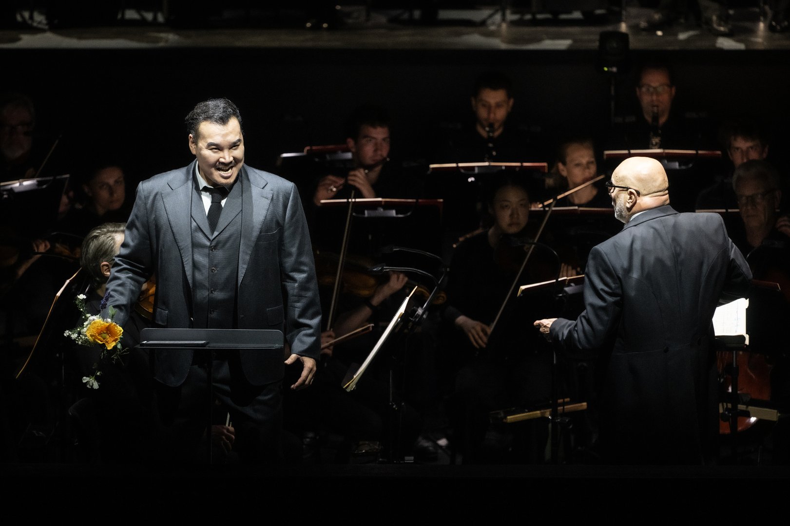 Auf der Bühne steht Young Woo Kim (Ernani) im Vordergrund und singt ausdrucksstark neben einem Notenpult, in der Hand eine Blume. Rechts von ihm dirigiert Giuliano Carella (musikalische Leitung) mit leicht erhobenem Arm und wendet sich dem Orchester zu. Im Hintergrund ist das Gürzenich Orchester zu sehen, dessen Musiker:innen konzentriert ihre Instrumente spielen.