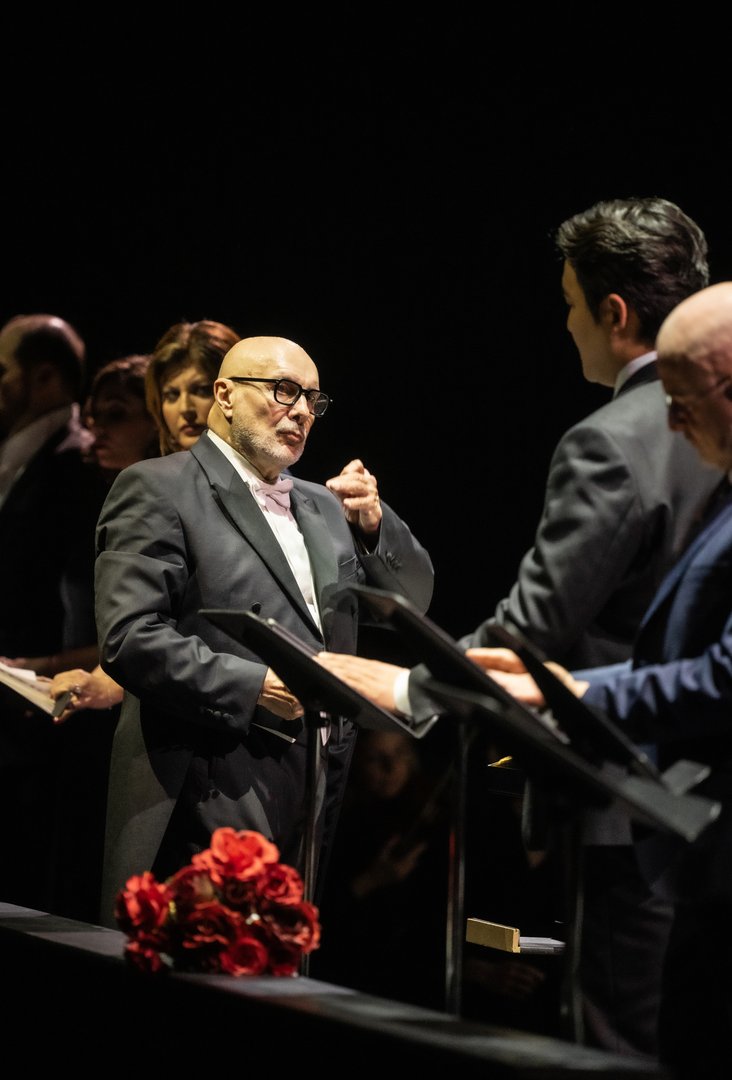 Im Vordergrund steht John Heuzenroeder (Don Riccardo) am Notenpult und singt mit konzentrierter Mimik. Rechts von ihm sind Insik Choi (Don Carlos) und Adrian Sâmpetrean (Don Ruy Gómez de Silva) zu sehen, die ebenfalls singen. Im Hintergrund dirigiert Giuliano Carella (musikalische Leitung) das Ensemble, während Marta Torbidoni (Elvira) und Alina König Rannenberg (Giovanna) hinter ihm stehen und am Geschehen beteiligt sind.