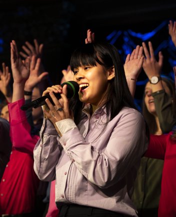 A young singer from the Kölner Jugendchor St. Stephan performs in the foreground with a microphone, wearing a light purple blouse and dark trousers. Behind her, other members of the choir stand in various colorful outfits, including red and green tops, singing along with raised hands and visible enthusiasm. The scene is illuminated by stage lighting, creating an energetic and festive concert atmosphere.