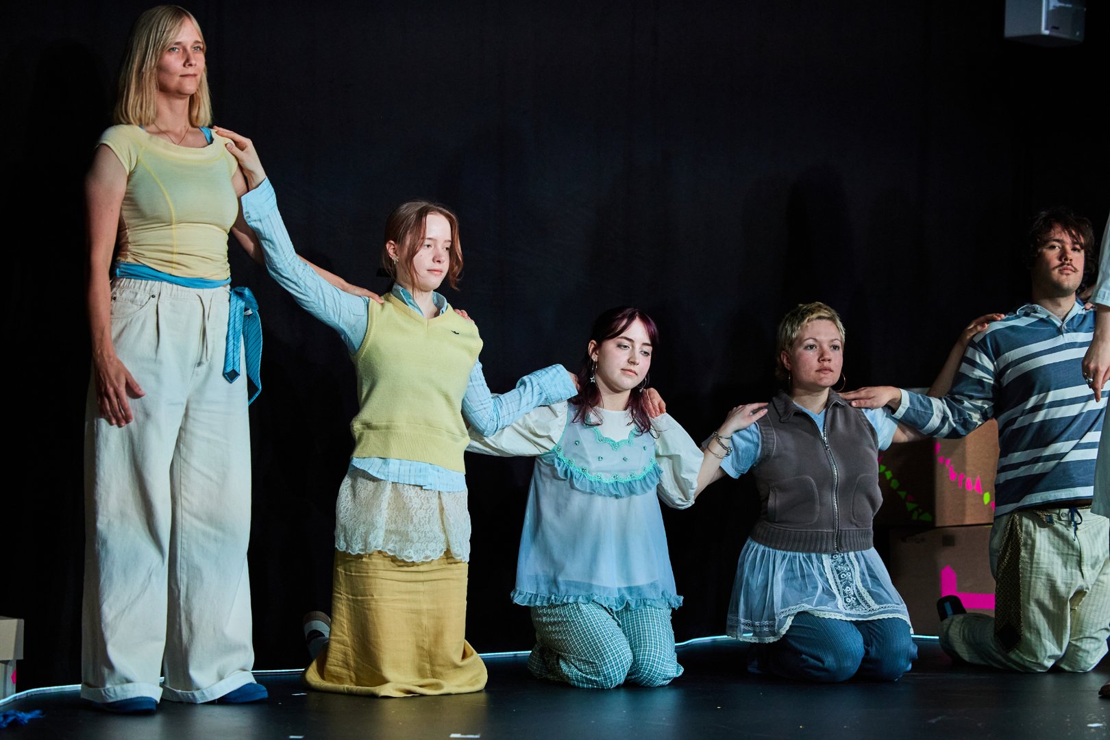 Szene aus When I find myself in Times of Trouble des Spielclubs Opera, Song and Scene. Fünf Mitwirkende bilden auf einer dunkel gehaltenen Bühne eine zusammenhängende Reihe. Die linke Person steht, die nächste steht auf den Knien, die nächste kniet, die nächste sitzt und die letzte steht auf ihren Knien. Alle sind durch aufgelegte Hände miteinander verbunden. Jede Person berührt die Schultern der benachbarten Person, sodass eine Kette aus Körperkontakt und gegenseitiger Unterstützung entsteht. Die Gesichter wirken ruhig und konzentriert, die Blicke sind nach vorn oder leicht in die Ferne gerichtet. Die Kleidung in hellen Blau-, Grün- und Cremetönen verleiht der Gruppe ein harmonisches Erscheinungsbild. Im Hintergrund sind Teile des Bühnenbilds mit Kisten und farbigen Markierungen zu erkennen, während ein Lichtband am Bühnenrand die Szene sanft umrahmt.