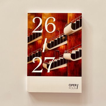 Das neue Spielzeitheft 2026/27 der Oper Köln ist vor einem hellen Hintergrund abgebildet. Auf dem Cover sind die Balkone des Zuschauerraums am Offenbachplatz mit warmen Holzvertäfelungen und geschwungenen, übereinander angeordneten Strukturen zu sehen. Groß steht „26 / 27“ auf dem Titel, unten befindet sich das Logo der Oper Köln.