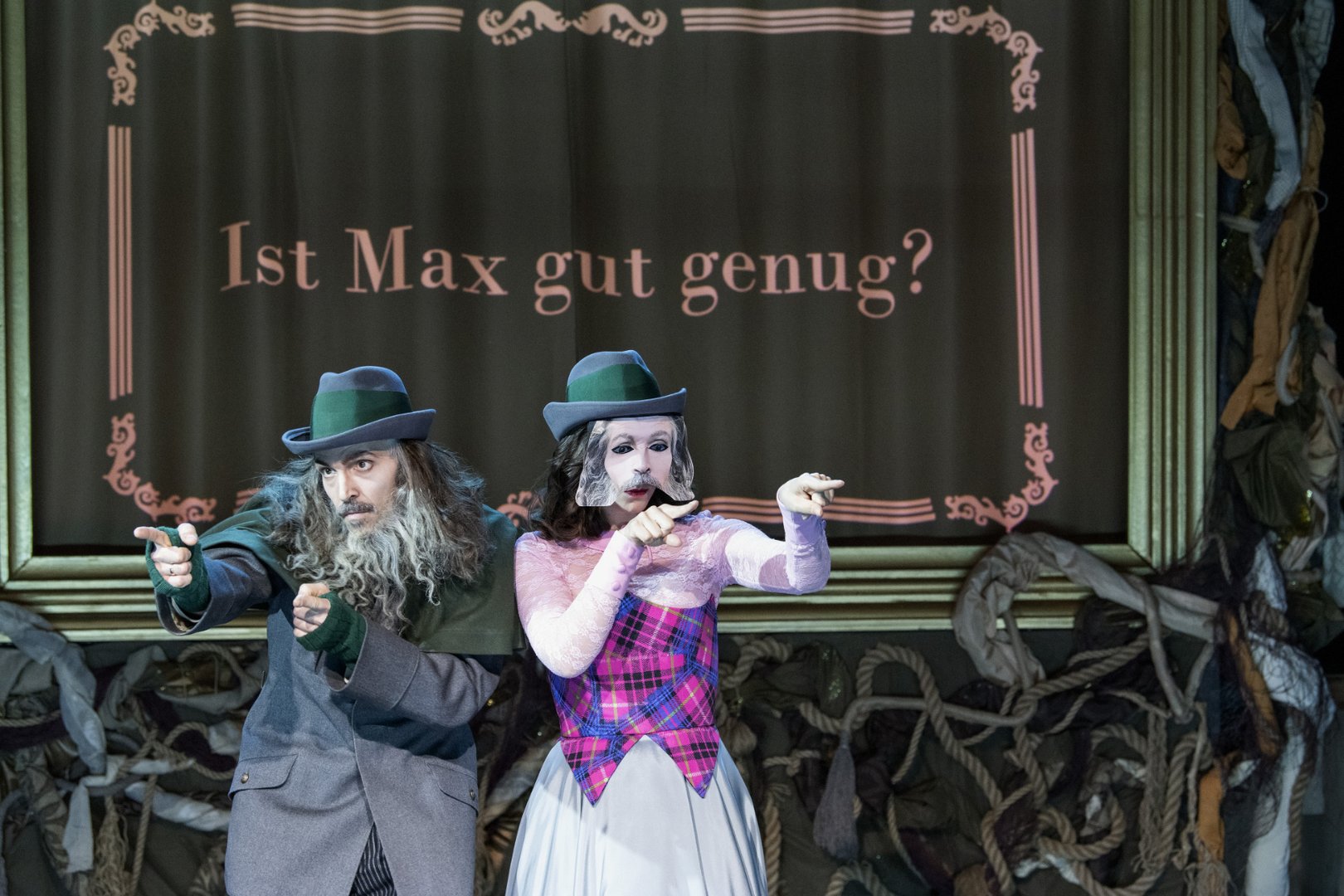 Erbförster Kuno und Ännchen aus der Oper „Freikugeln“ stehen nebeneinander auf der Bühne vor einem dekorativen Vorhang, auf dem der Satz „Ist Max gut genug?“ zu lesen ist. Beide tragen grüne Hüte; Kaspar ist in dunklere, jagdliche Kleidung mit langem Bart gekleidet, während Ännchen ein farbenfroheres Kostüm mit pink kariertem Oberteil und heller Rockpartie trägt.
Beide Figuren zeigen gleichzeitig eine Gebärde: Ihre Arme sind nach vorne gerichtet, die Hände zu einer Art „Zeigegeste“ geformt, wobei die Finger ausgestreckt sind und in dieselbe Richtung weisen. Die Haltung wirkt synchron und bewusst gesetzt, als würden sie gemeinsam auf etwas oder jemanden aufmerksam machen. Ihre Blicke folgen der Richtung der Hände, wodurch die Szene eine klare, fokussierte Spannung erhält. Im unteren Bühnenbereich sind dekorative Elemente wie Seile und Stoffe sichtbar.
