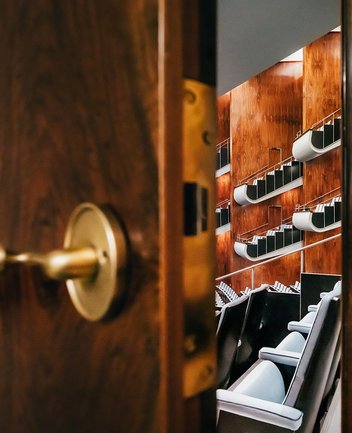 Durch eine leicht geöffnete Tür fällt der Blick in den Zuschauerraum der Oper Köln am Offenbachplatz. Im Vordergrund ist die Tür mit Griff zu sehen, dahinter öffnen sich gestaffelte Sitzreihen und Ränge, die die Tiefe und Architektur des Saals erahnen lassen.