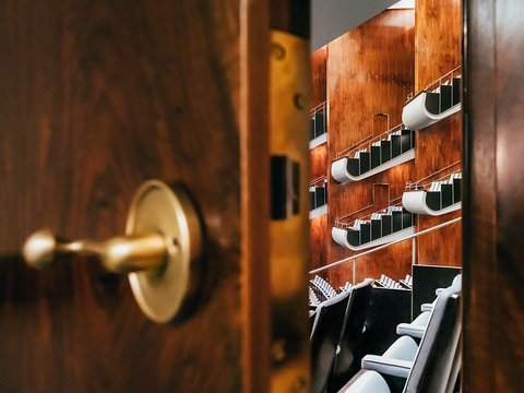 Durch eine leicht geöffnete Tür fällt der Blick in den Zuschauerraum der Oper Köln am Offenbachplatz. Im Vordergrund ist die Tür mit Griff zu sehen, dahinter öffnen sich gestaffelte Sitzreihen und Ränge, die die Tiefe und Architektur des Saals erahnen lassen.
