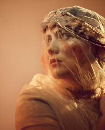 Nahaufnahme einer jungen Braut mit cineastischer Bildwirkung und warmer Lichtstimmung: Eine junge Frau mit zarten Gesichtszügen trägt einen filigranen, transparenten Schleier, der mit Perlen und feinen Verzierungen am Kopfstück geschmückt ist. Ihr Blick ist leicht nach oben links gerichtet, als würde sie in die Ferne oder eine ungewisse Zukunft schauen. Der Schleier legt sich sanft über ihr Gesicht und verleiht der Szene eine träumerische, fast entrückte Atmosphäre. Ihr Ausdruck vereint Sehnsucht und Entschlossenheit, während die warmen Farbtöne und die feinen Stoffstrukturen eine intime, poetische Bildwirkung erzeugen. Die Szene evoziert die Figur der Marie aus Bedřich Smetanas Oper Die verkaufte Braut.