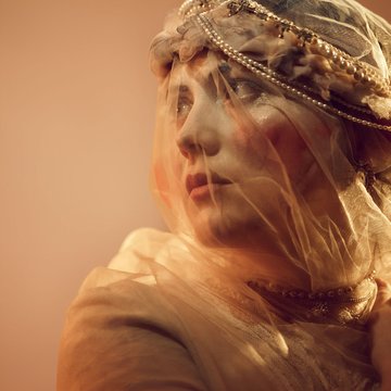 Close-up of a young bride with a cinematic look and warm lighting: A young woman with delicate features wears a fine, transparent veil adorned with pearls and intricate embellishments at the headpiece. Her gaze is directed slightly upward to the left, as if looking into the distance or toward an uncertain future. The veil gently drapes over her face, lending the scene a dreamlike, almost ethereal atmosphere. Her expression combines longing and determination, while the warm tones and delicate textures of the fabric create an intimate, poetic visual effect. The scene evokes the figure of Marie from Bedřich Smetana’s opera The Bartered Bride.