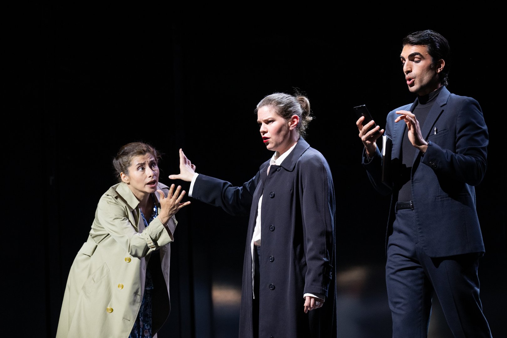 Drei Figuren aus der Oper Picture a day like this stehen in einer dramatisch ausgeleuchteten Bühnenszene nebeneinander. Links richtet die Woman in einem hellen Trenchcoat eine eindringliche Geste an die beiden anderen Figuren. In der Mitte hebt Lover 1 / Composer die Hand abwehrend oder beschwichtigend, während rechts Lover 2 / Composer’s Assistant in dunklem Anzug auf ein Smartphone blickt und gleichzeitig spricht oder singt. Der schwarze Bühnenraum und das konzentrierte Licht lenken den Fokus ganz auf die ausdrucksstarken Gesten und die spannungsvolle Interaktion der Figuren.