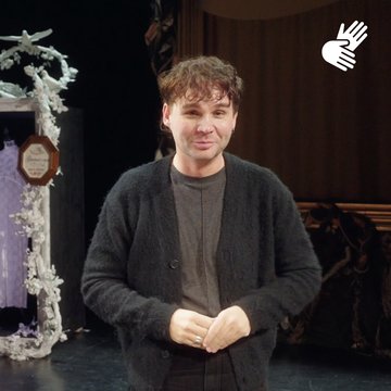 Rafael-Evitan Grombelka steht frontal auf der Probebühne zur Produktion Freikugeln und blickt lächelnd in die Kamera. Er trägt ein dunkles T-Shirt unter einer schwarzen Strickjacke, die Hände locker vor dem Körper gefaltet. Im Hintergrund ist ein Teil des Bühnenbilds zu sehen: links ein dekoratives, helles Gestell mit floralen Elementen und einem kleinen Schild, rechts ein dunkler Vorhang. Oben rechts im Bild befindet sich ein eingeblendetes Symbol für Deutsche Gebärdensprache (DGS) – zwei Hände mit ausgestreckten Fingern, die sich kreuzen. Die Szene ist warm ausgeleuchtet und vermittelt eine konzentrierte, aber entspannte Probenatmosphäre.