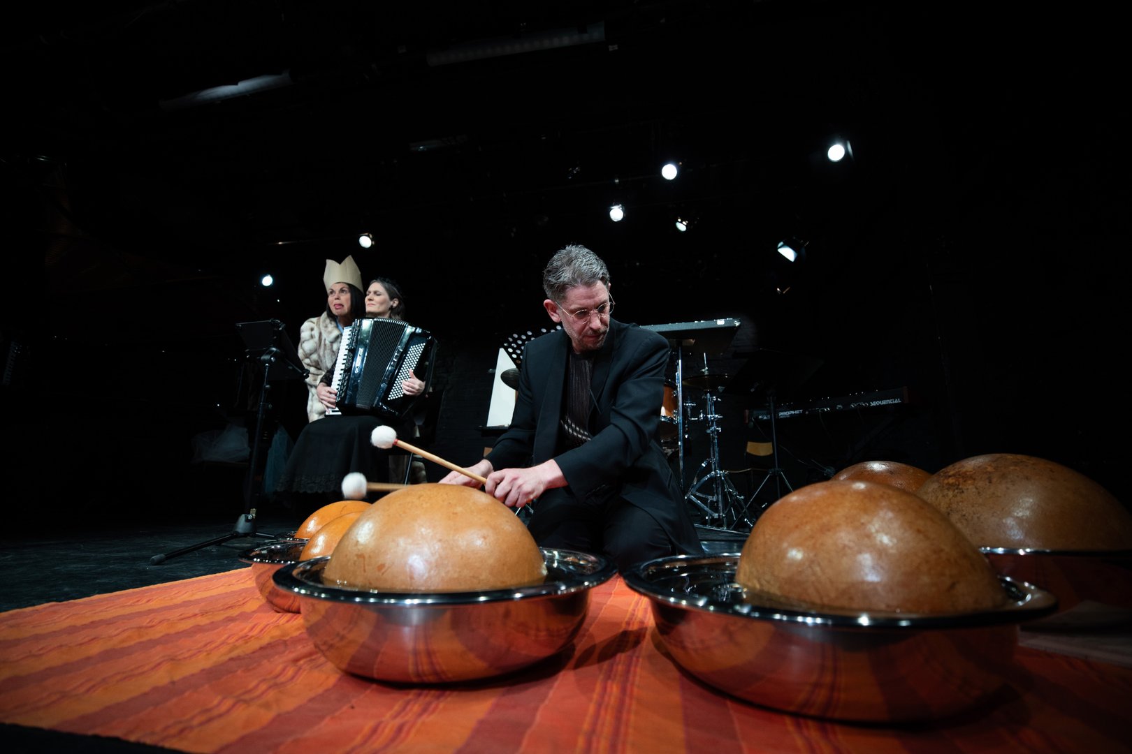 Kurt Fuhrmann sitzt auf einer Bühne und spielt mit Schlägeln auf halbkugelförmige Percussion-Instrumente in Metallschalen. Im Hintergrund sind KS Dalia Schaechter und Dorrit Bauerecker zu sehen. Dorrit Bauerecker mit Akkordeon und Dalia Schaechter mit auffälligem Kostüm und Kopfschmuck. Die Szene ist dunkel ausgeleuchtet und wirkt wie eine experimentelle Theater- oder Musikperformance.