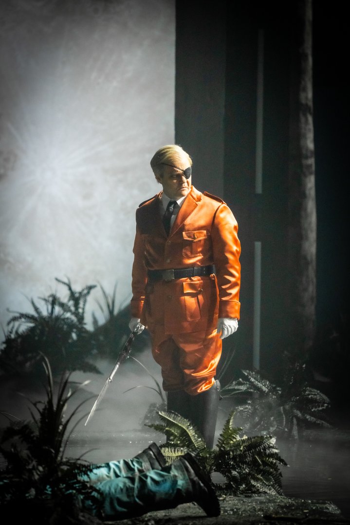 Auf einer nebligen Bühne steht Wotan in einem auffälligen orangefarbenen Anzug mit Augenklappe und hält ein Schwert locker in der Hand. Sein Blick ist ernst und nach unten gerichtet, wo eine am Boden liegende Figur angedeutet ist. Umgeben von Pflanzen und gedämpftem Licht entsteht eine spannungsgeladene Szene aus Die Walküre.