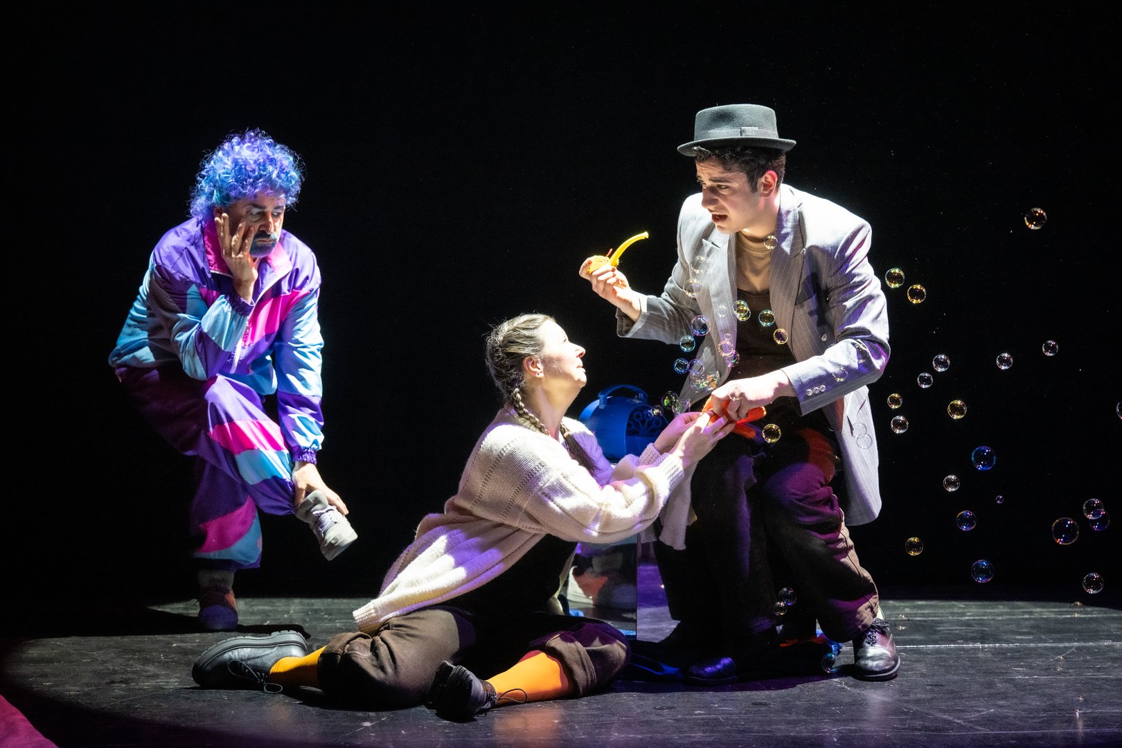 Szene aus der Kinderoper Max und Moritz: Auf der Bühne sitzen Frau Gagackel in dunkler Latzhose und weißer Strickjacke und Henriette, das Huhn / Herr Guru / Dr. Gehirn / Belinda, die Tarantel in dunkler Latzhose, gestreiftem Shirt und Hut. Sie lassen zahlreiche Seifenblasen in die Luft steigen. Links daneben beobachtet Trouble die Szene in einem bunten Trainingsanzug (Lila, Pink und Türkis) mit blauer Lockenperücke. Er wirkt gelangweilt. Die dunkle Bühne wird von warmem Licht und schwebenden Seifenblasen geprägt.