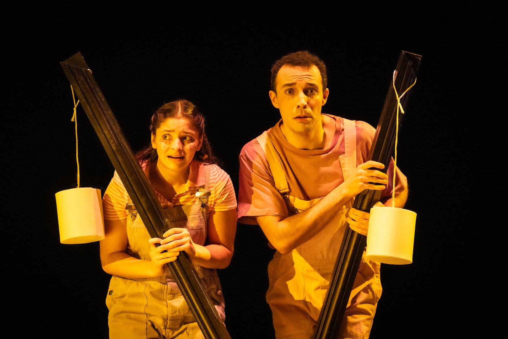 Szene aus der Kinderoper Max und Moritz: Auf einer dunklen Bühne stehen Max und Moritz eng beieinander und schauen erschrocken ins Publikum. Beide tragen hellrosa Latzhosen über rosafarbenen T-Shirts. Sie halten jeweils ein langes, schwarzes Brett wie ein Schutzschild vor sich; daran hängt an einer Schnur eine weiße Toilettenpapierrolle. Die Szene wirkt wie ein Moment des Lauschens oder Versteckens im Scheinwerferlicht.