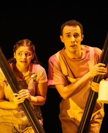 Szene aus der Kinderoper Max und Moritz: Auf einer dunklen Bühne stehen Max und Moritz eng beieinander und schauen erschrocken ins Publikum. Beide tragen hellrosa Latzhosen über rosafarbenen T-Shirts. Sie halten jeweils ein langes, schwarzes Brett wie ein Schutzschild vor sich; daran hängt an einer Schnur eine weiße Toilettenpapierrolle. Die Szene wirkt wie ein Moment des Lauschens oder Versteckens im Scheinwerferlicht.