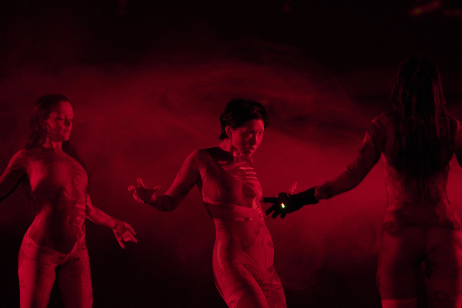 Drei Tänzerinnen bewegen sich in einer rot ausgeleuchteten Bühne vor einer wirbelnden, abstrakten Projektion. Die Person in der Mitte steht leicht nach hinten geneigt, die Arme seitlich geöffnet. Ihre Hände sind angespannt und ausdrucksstark geformt: Finger gespreizt und leicht gekrümmt, als würden sie etwas Unsichtbares halten oder formen. Links und rechts greifen die anderen Performerinnen diese Körpersprache auf – ihre Hände hängen oder öffnen sich in ähnlichen, kontrollierten Gesten, mit weichen, aber präzisen Fingerstellungen. Die Handbewegungen wirken wie ein gemeinsames, stilles Vokabular und verstärken die intensive, beinahe magnetische Atmosphäre der Szene.