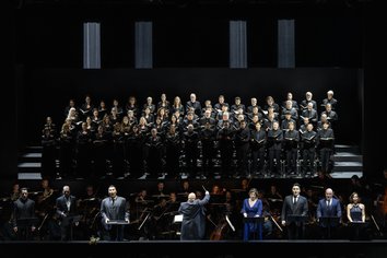 Auf der Bühne ist eine große Ensembleszene zu sehen: Im Vordergrund stehen Ferhat Baday (Jago), Adrian Sâmpetrean (Don Ruy Gómez de Silva), Young Woo Kim (Ernani), Marta Torbidoni (Elvira), Insik Choi (Don Carlos), John Heuzenroeder (Don Riccardo) und Alina König Rannenberg (Giovanna) an Notenpulten und singen. In der Mitte dirigiert Giuliano Carella (musikalische Leitung) das Gürzenich Orchester, das im Orchestergraben sitzt. Im Hintergrund ist der Chor der Oper Köln auf mehreren Podesten aufgestellt und singt geschlossen als großes Ensemble.