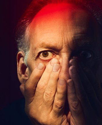 Nahaufnahme eines älteren Mannes mit cineastischer Bildwirkung und warmer Lichtstimmung: Das Gesicht ist frontal zu sehen, der Blick intensiv und weit geöffnet; die Augen wirken erschrocken und angespannt. Die rechte Gesichtshälfte liegt im Schatten. Beide Hände sind vor das Gesicht gelegt, die Finger bedecken Nase, Mund und Wangen – als wolle er sich schützen oder seine Wahrnehmung abschirmen. Die Haut zeigt feine Linien und Falten und glänzt im starken Bühnenlicht. Dramatisches Licht in intensiven Rot- und Blautönen fällt von oben und der Seite auf das Gesicht, wodurch starke Kontraste entstehen und die psychische Anspannung der Figur hervorgehoben wird. Der dunkle Hintergrund tritt vollständig zurück, sodass der Fokus ganz auf dem Ausdruck von Angst, Schuld und innerer Unruhe liegt. Die Szene evoziert die Figur des Macbeth aus Giuseppe Verdis Oper Macbeth.