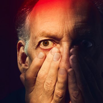Close-up of an older man with a cinematic look and warm lighting: The face is shown frontally, the gaze intense and wide open; the eyes appear startled and tense. The right side of the face is cast in shadow. Both hands are raised in front of the face, the fingers covering the nose, mouth, and cheeks—as if to protect himself or block out his perception. The skin shows fine lines and wrinkles and glistens under strong stage lighting. Dramatic light in intense red and blue tones falls from above and the side onto the face, creating strong contrasts and emphasizing the character’s psychological tension. The dark background recedes completely, focusing attention entirely on the expression of fear, guilt, and inner turmoil. The scene evokes the character of Macbeth from Giuseppe Verdi’s opera Macbeth.