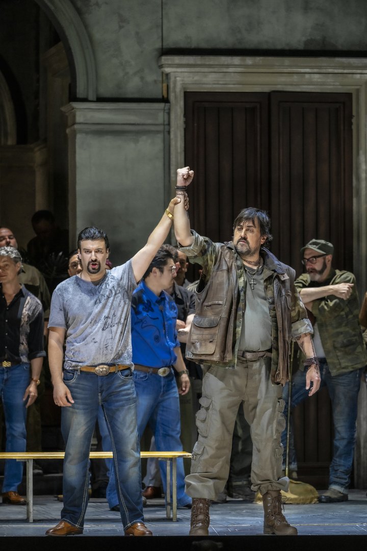 Auf einer Opernbühne stehen Argirio und Orbazzano aus Rossinis Tancredi im Mittelpunkt einer Gruppenszene. Argirio trägt Jeans und ein graues T-Shirt, Orbazzano eine militärisch anmutende Kleidung mit Weste und Stiefeln. Gemeinsam heben sie die verschränkten Arme als Zeichen von Zusammenhalt und Macht. Hinter ihnen versammelt sich eine Gruppe weiterer Personen. Das Bühnenbild zeigt die Fassade einer großen Villa mit hohen Türen und verleiht der Szene die Atmosphäre eines von rivalisierenden Clans geprägten Milieus. Die Inszenierung verlegt die Handlung in die Welt eines Drogenkartells und betont Themen wie Loyalität, Gewalt und Machtstrukturen.