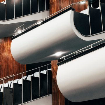 Blick in den Zuschauerraum der Oper Köln am Offenbachplatz mit modernen, geschwungenen Balkonlogen. Die Logen zeichnen sich durch glatte, helle Oberflächen und dunkle Geländer aus und sind über mehrere Ebenen angeordnet. Im Hintergrund sind warme Holzvertäfelungen sowie darunterliegende Sitzreihen zu sehen, wodurch ein eleganter und zeitgenössischer Raumeindruck entsteht.