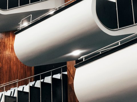 Blick in den Zuschauerraum der Oper Köln am Offenbachplatz mit modernen, geschwungenen Balkonlogen. Die Logen zeichnen sich durch glatte, helle Oberflächen und dunkle Geländer aus und sind über mehrere Ebenen angeordnet. Im Hintergrund sind warme Holzvertäfelungen sowie darunterliegende Sitzreihen zu sehen, wodurch ein eleganter und zeitgenössischer Raumeindruck entsteht.