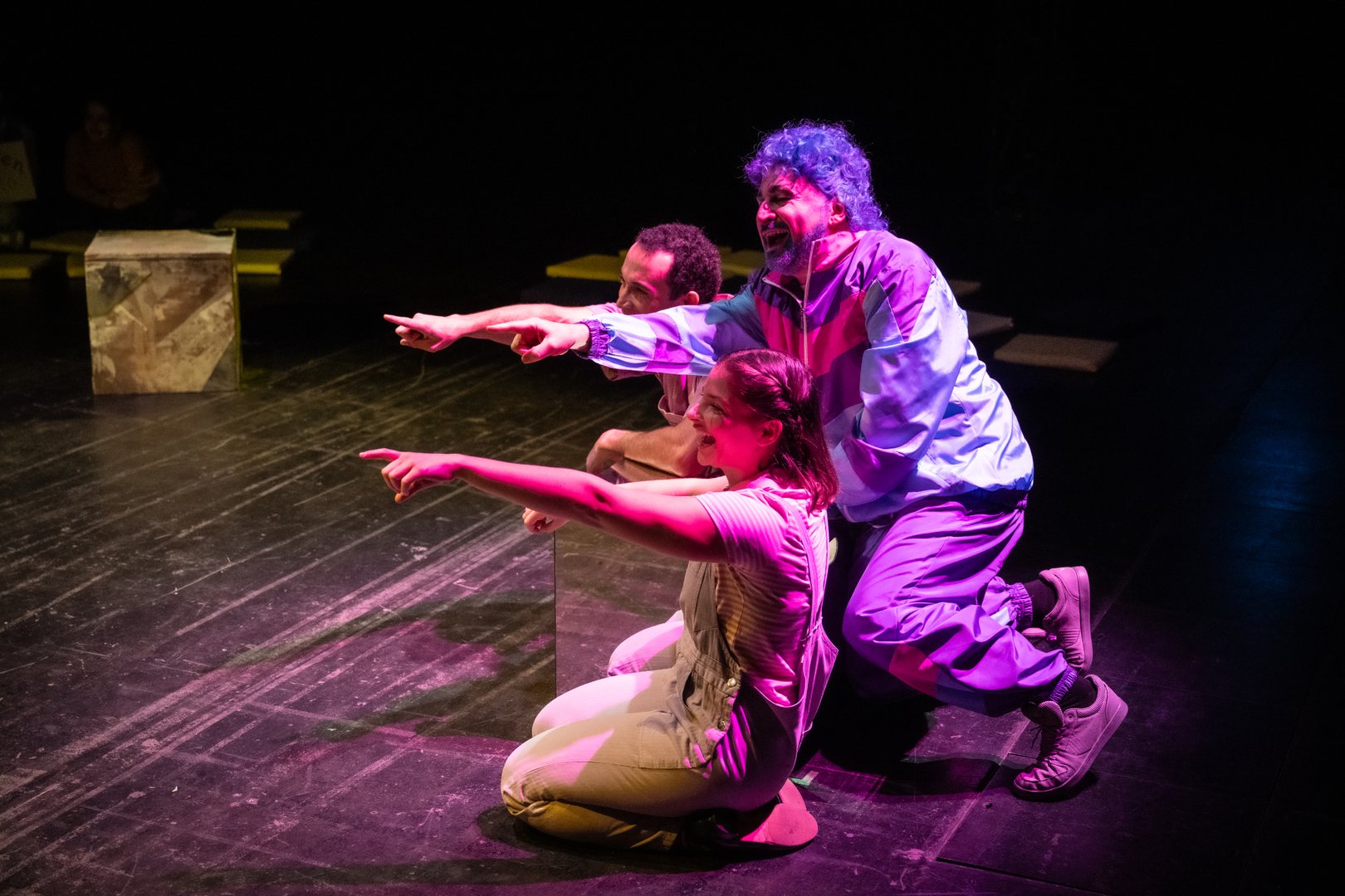 Szene aus der Kinderoper Max und Moritz: Auf der Bühne hocken Max, Moritz und Trouble im Vordergrund auf dem Boden. Max und Moritz tragen beide hellrosa Latzhosen und lila T-Shirts. Hinter ihnen steht Trouble in einem bunten Trainingsanzug (Lila, Pink und Türkis) mit blauer Lockenperücke. Alle drei zeigen links aus dem Bildausschnitt und lachen Jemanden oder etwas mit erhobenen Zeigefindern aus. Die dunkle Bühne wird von rosafarbenem Licht geprägt.