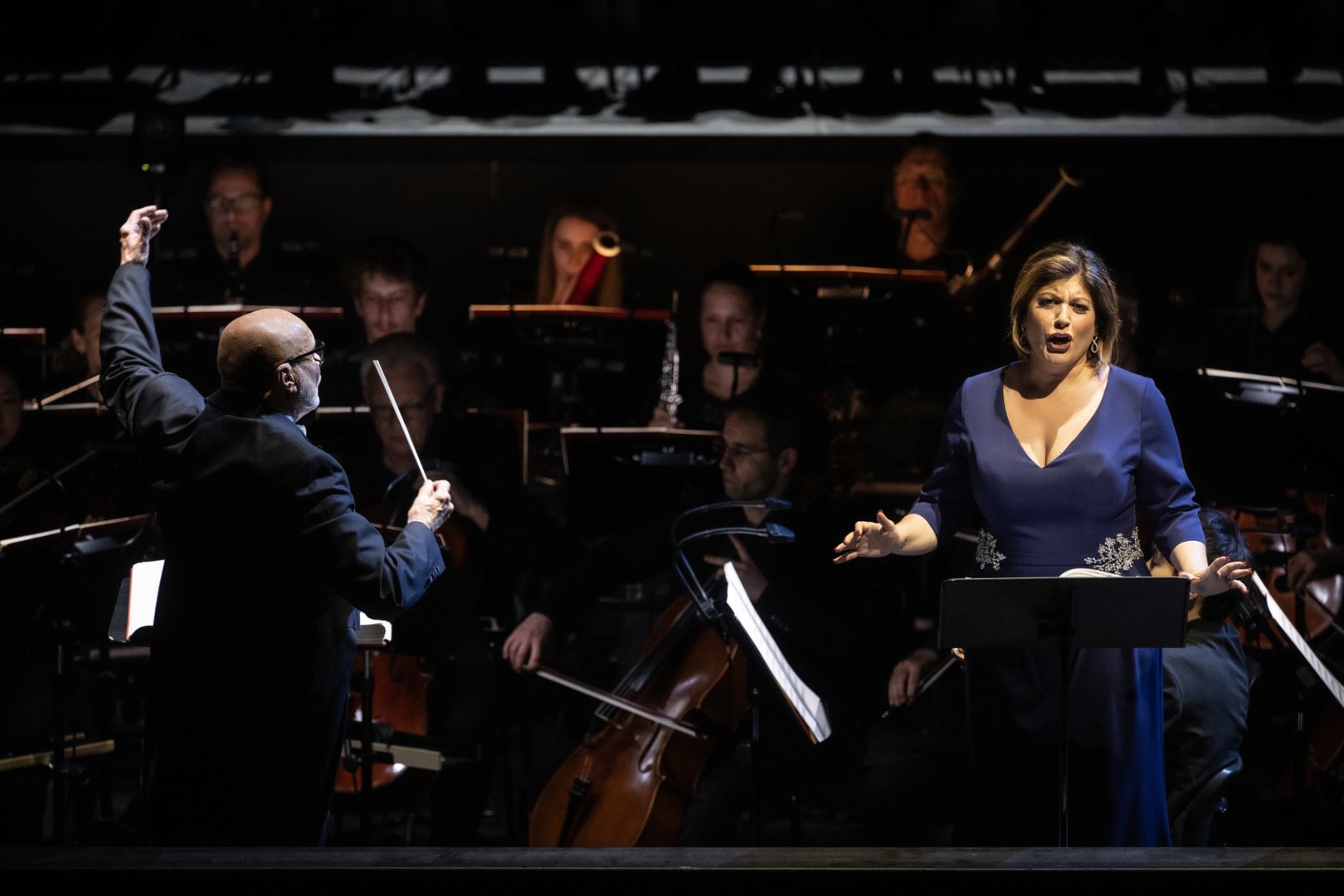 Auf der Bühne dirigiert Giuliano Carella (musikalische Leitung) mit erhobenem Taktstock das Gürzenich Orchester. Rechts von ihm steht Marta Torbidoni (Elvira) in einem blauen Kleid am Notenpult und singt mit ausdrucksstarker Gestik. Im Hintergrund sind die Musiker*innen des Orchesters zu sehen, die konzentriert ihre Instrumente spielen.