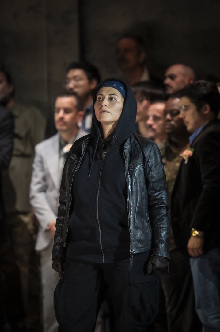 Tancredi aus Rossinis Tancredi steht im Mittelpunkt einer Gruppe von Menschen. Die Figur trägt dunkle Kleidung mit Kapuzenjacke, Lederjacke, Handschuhen und einem blauen Kopftuch. Mit ernstem, konzentriertem Blick schaut Tancredi nach oben, während die Personen im Hintergrund unscharf bleiben. Das Bühnenlicht lenkt die Aufmerksamkeit auf die isoliert wirkende Figur und unterstreicht ihre innere Anspannung. Die Szene vermittelt Entschlossenheit, Einsamkeit und die ständige Gefahr eines Lebens im Verborgenen innerhalb der von rivalisierenden Clans geprägten Welt dieser Inszenierung.
